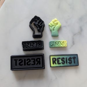 May include: A set of five cookie cutters, two are black and shaped like a fist, one is black and says "RESIST" in reverse, one is green and yellow and says "RESIST", and one is green and yellow and shaped like a fist.