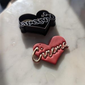 May include: Two heart-shaped cookie cutters, one black and one red. The black cutter is inscribed with the word "mommy" and the red cutter is inscribed with the word "cinema".