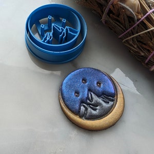 May include: A blue plastic cookie cutter with a mountain range design next to a decorated cookie. The cookie has a blue and gold color scheme with a mountain range design and three small holes.