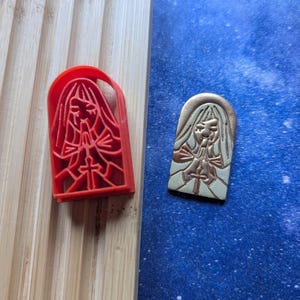 Puede incluir: Dos colgantes de cerámica, uno rojo y otro blanco, con una imagen en relieve de una persona rezando con una cruz. Ambos colgantes son rectangulares y tienen una parte superior ligeramente arqueada.