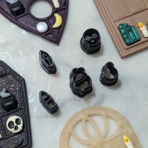 Puede incluir: Un juego de cortadores de galletas de plástico negro con forma de ataúd, luna creciente, caldero, fantasma y sombrero de bruja. Los cortadores están sobre una superficie blanca.
