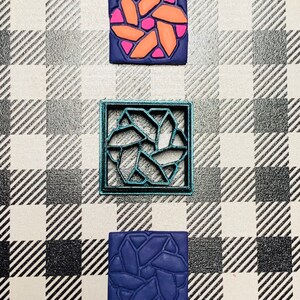May include: Three square-shaped polymer clay tiles with a geometric design. The top tile is pink and orange, the middle tile is teal, and the bottom tile is blue.