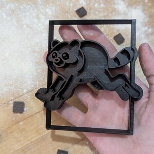 May include: A black plastic cookie cutter featuring a stylized raccoon design enclosed within a rectangular frame. The raccoon is depicted in a leaping or running pose with its tail extended, showcasing detailed facial features and a striped tail.
