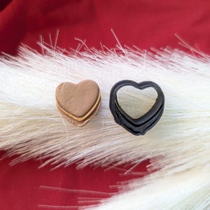 Puede incluir: Dos objetos en forma de corazón. Uno es un cortador de galletas marrón claro en capas. El otro es un cortador de galletas negro y estriado. Ambos están sobre una superficie blanca y esponjosa sobre un fondo rojo. Ideales para hornear y manualidades.