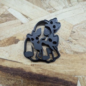 May include: A black plastic cookie cutter featuring a stylized design of two small cat-like figures with large eyes and pointed ears, positioned together with a small star shape above them, all contained within a single continuous frame.
