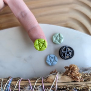 May include: A set of five small, round, clay pentagram charms. The charms are in various colors, including green, blue, and black. The charms are on a white surface.
