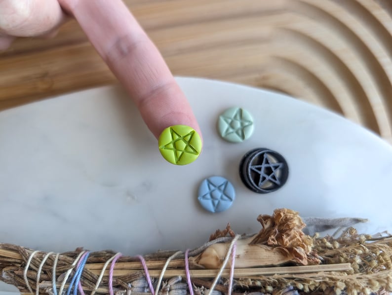 May include: A set of five small, round, clay pentagram charms. The charms are in various colours, including green, blue, and black. The charms are on a white surface.