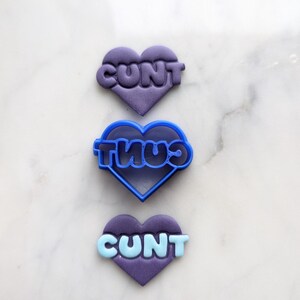 May include: Three heart-shaped cookie cutters, two purple and one blue. The cutters are shaped like hearts and have the word "CUNT" written on them in a bubble-like font.