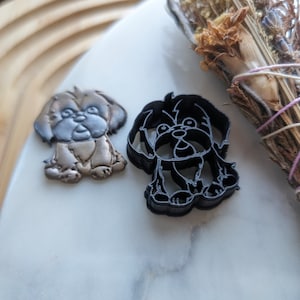 Puede incluir: Dos cortadores de galletas con forma de perro, uno marrón y otro negro. El cortador de galletas marrón es una representación realista de un perro, mientras que el cortador de galletas negro es una versión más estilizada.