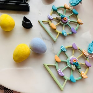 Puede incluir: Un par de pendientes caprichosos de rueda de la fortuna con detalles en colores pastel. Los pendientes presentan un marco verde claro, asientos coloridos y una parte superior en forma de nube azul. También se ven varios huevos de colores y un cortador en forma de rueda de la fortuna.