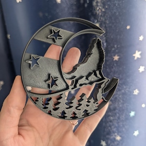 Puede incluir: Un cortador de galletas de plástico negro en forma de media luna con un lobo aullando a la luna. Hay tres estrellas y un bosque de pinos en el fondo.