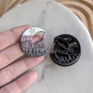 Puede incluir: Dos cortadores de galletas en forma de media luna, uno negro y otro con un degradado de blanco, rosa y marrón. Ambos cortadores presentan un lobo aullando a la luna y una escena de bosque con estrellas.