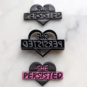 Puede incluir: Tres cortadores de galletas en forma de corazón negro con el texto "She Persisted" en blanco. El cortador superior tiene el texto en línea recta, el cortador del medio tiene el texto al revés y el cortador inferior tiene el texto en línea recta con una banda rosa.