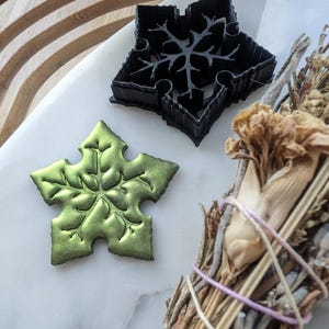 Könnte beinhalten: Ein grüner Schneeflocken-Ausstecher und ein schwarzer Schneeflocken-Ausstecher aus Kunststoff auf einer weißen Oberfläche. Im Hintergrund befindet sich ein Bündel getrockneter Kräuter.