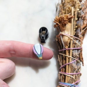 Puede incluir: Un pequeño amuleto de globo aerostático rosa y blanco con una cesta gris y un detalle dorado. El amuleto está en un dedo.