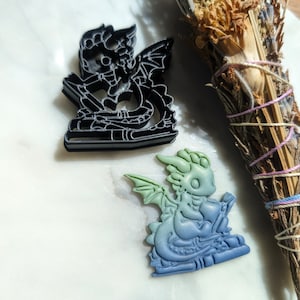 Puede incluir: Dos cortadores de galletas de dragón, uno negro y otro verde y azul. El cortador negro es una silueta de un dragón leyendo un libro. El cortador verde y azul es un dragón 3D leyendo un libro.