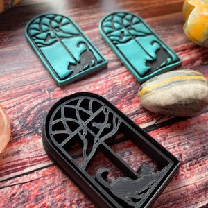 May include: Three cookie cutters in the shape of a window with a cat inside. The cutters are made of black plastic and are on a wooden surface. The two smaller cutters are teal blue.