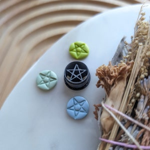 May include: A black pentagram charm with a five-pointed star design. The charm is surrounded by four smaller charms, two green, one blue, and one light blue, each with a five-pointed star design.