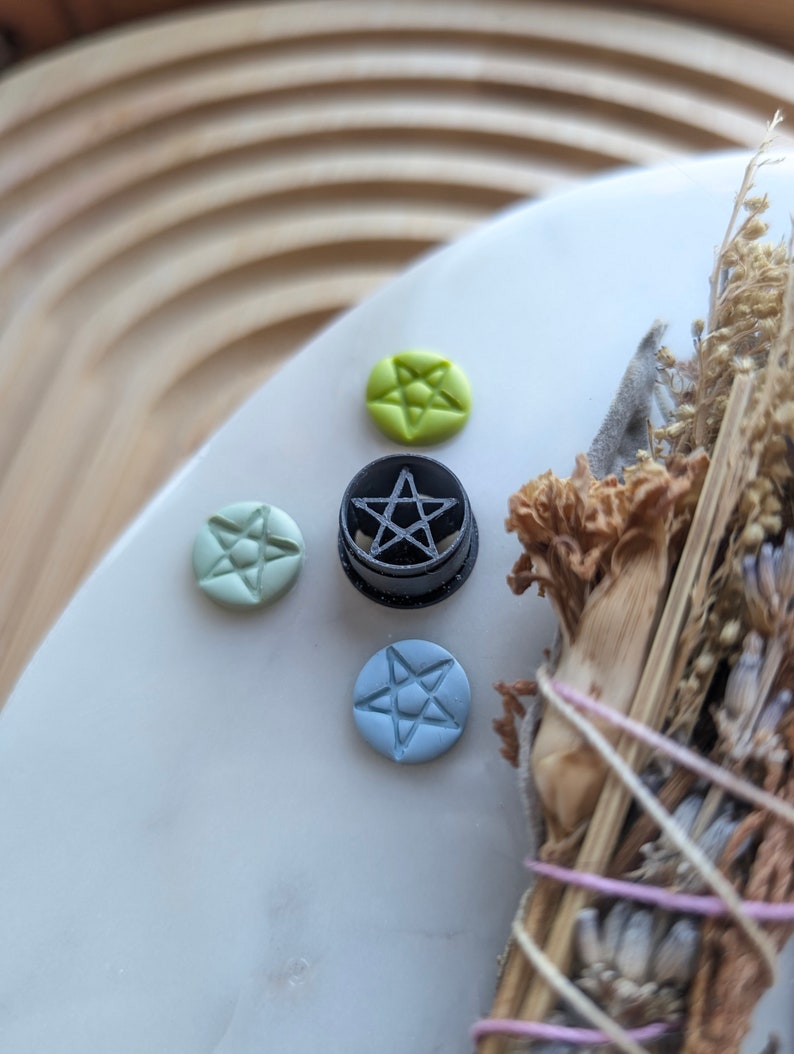 May include: A black pentagram charm with a five-pointed star design. The charm is surrounded by four smaller charms, two green, one blue, and one light blue, each with a five-pointed star design.