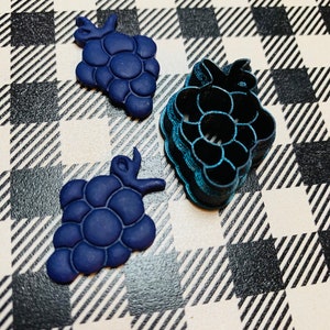 Puede incluir: Tres cortadores de galletas con forma de uva, dos son azules y uno es turquesa. Los cortadores azules están hechos de arcilla polimérica y el cortador turquesa está hecho de metal.