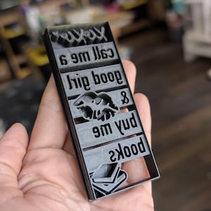 May include: A black rectangular stamp with the words "yayay", "6 9m lls3", "hip boop", "&", "9m yud", and "skood" in a stylized font. The stamp also features an image of a pair of animals and an image of a book.
