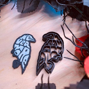Puede incluir: Dos cortadores de galletas de cuervo negro, uno con las palabras "Nevermore" escritas en él, y el otro con un diseño más detallado. Los cortadores de galletas están sobre una superficie de madera.