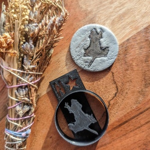 May include: A bundle of dried herbs and lavender tied with string, next to a grey, circular coaster with a lizard silhouette. A black, circular object with a lizard silhouette and a rectangular object with a star are also present.
