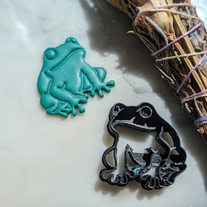 May include: Two frog figurines, one green and one black, on a white surface. The green frog is sitting with its legs bent, while the black frog is sitting with its legs stretched out.
