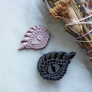 Puede incluir: Dos moldes de arcilla polimérica en forma de ojo de dragón. Uno es de color gris oscuro y el otro es de color oro rosa.