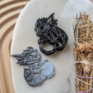 Puede incluir: Dos cortadores de galletas negros con forma de dragones. Un dragón sostiene una olla. El dragón de la izquierda es de color azul claro.