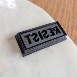 May include: A black rectangular stamp with the word "RESIST" in a bold, sans-serif font. The stamp is sitting on a white marble surface, with a wooden background.
