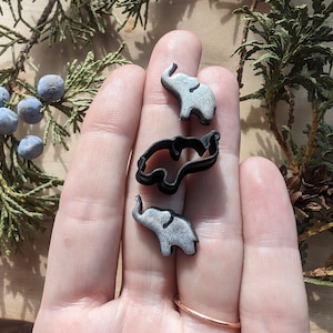 May include: Three silver-colored elephant cookie cutters, two are standing and one is lying down. The cookie cutters are on a person's hand.
