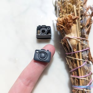May include: A small black camera charm with a detailed lens and a textured surface.