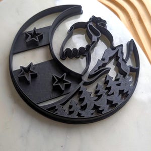 Puede incluir: Cortador de galletas de plástico negro con una escena de montaña con un oso, una luna, estrellas y árboles. El cortador tiene forma de media luna y está diseñado para hornear. El diseño es detallado.