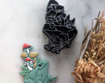 May include: Two frog cookie cutters, one black and one green. The black cutter is of a frog sitting on top of a mushroom. The green cutter is of a frog with a mushroom on its head.