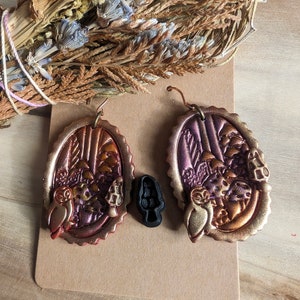 May include: A pair of owl themed earrings with a forest scene, featuring a brown, gold, and purple color scheme. The earrings are made of polymer clay and have a scalloped edge.