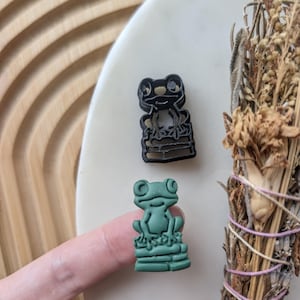 May include: Two frog figurines, one black and one green, sitting on a stack of bricks. The figurines are made of plastic and have a 3D design.