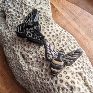 May include: Two bee-shaped pendants. One is solid black, the other has metallic silver wings and a black and gold striped body. They rest on a textured, off-white coral-like surface, with a wooden background. The pendants are likely for jewelry or crafts.