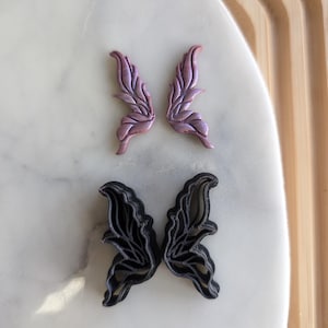 Puede incluir: Dos pares de alas de hada, un par es un cortador de galletas de plástico negro y el otro par es un recorte de arcilla polimérica rosa y morado.