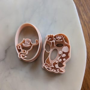 以下が含まれることがあります： 2つの薄いピンク色のプラスチック製クッキーカッター。1つは漫画のモンスターのデザインの楕円形です。もう1つは、別のモンスターデザインの、より大きく不規則な形状です。どちらも明るい色の表面にあります。