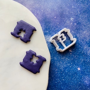 Puede incluir: Dos cortadores de galletas, uno blanco y otro morado, con forma de la letra "I" con un diseño decorativo.