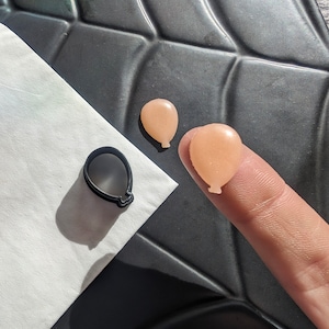 Puede incluir: Dos pequeños moldes de silicona en forma de globo de color rosa claro y un cortador de galletas de plástico negro en forma de globo.