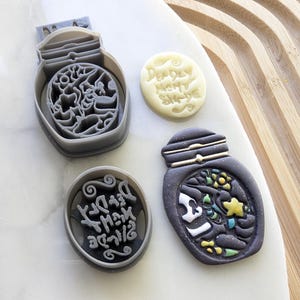 Puede incluir: Dos cortadores de galletas y dos galletas decoradas, todas con un frasco con tapa y el texto "Dream a little dream". Los cortadores de galletas son de plástico gris y las galletas están decoradas con glaseado negro, blanco, amarillo y verde.