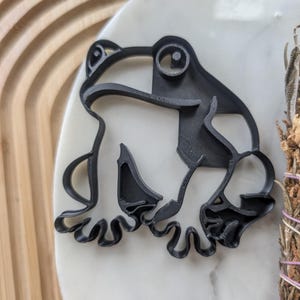 May include: Black plastic frog cookie cutter. The frog is in a sitting position with its legs outstretched.