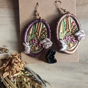 May include: A pair of oval-shaped earrings with a mushroom and bunny design. The earrings are made of polymer clay and have a metallic finish. The earrings are a mix of purple, green, and gold colors.