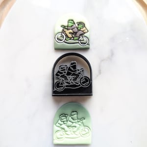 May include: Three green frogs riding a bicycle together. The frogs are wearing brown and yellow clothing. The bicycle is black with white wheels. The image is a close-up of a cookie cutter.