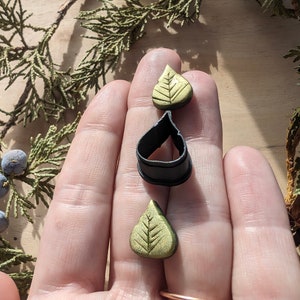 May include: Two gold leaf-shaped polymer clay charms with a black polymer clay ring. The charms have a textured leaf design.