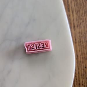 May include: A pink rectangular stamp with the word "RESIST" in a bold, sans-serif font. The stamp is set against a light-coloured, marble-like surface.