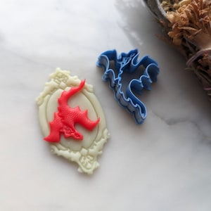 Puede incluir: Un cortador de galletas de dragón rojo con un marco blanco y un cortador de galletas de dragón azul. Ambos cortadores están sobre una superficie blanca.