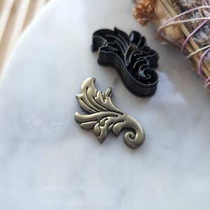 May include: Two decorative metal cookie cutters in the shape of a stylized leaf. The cutter on the left is gold, and the cutter on the right is black.